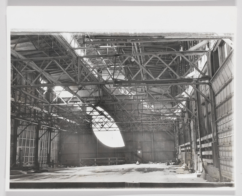 A cavernous, abandoned structure with exposed metal beams and a monumental, leaf-shaped aperture cut into the far wall and filling the space with sunlight.